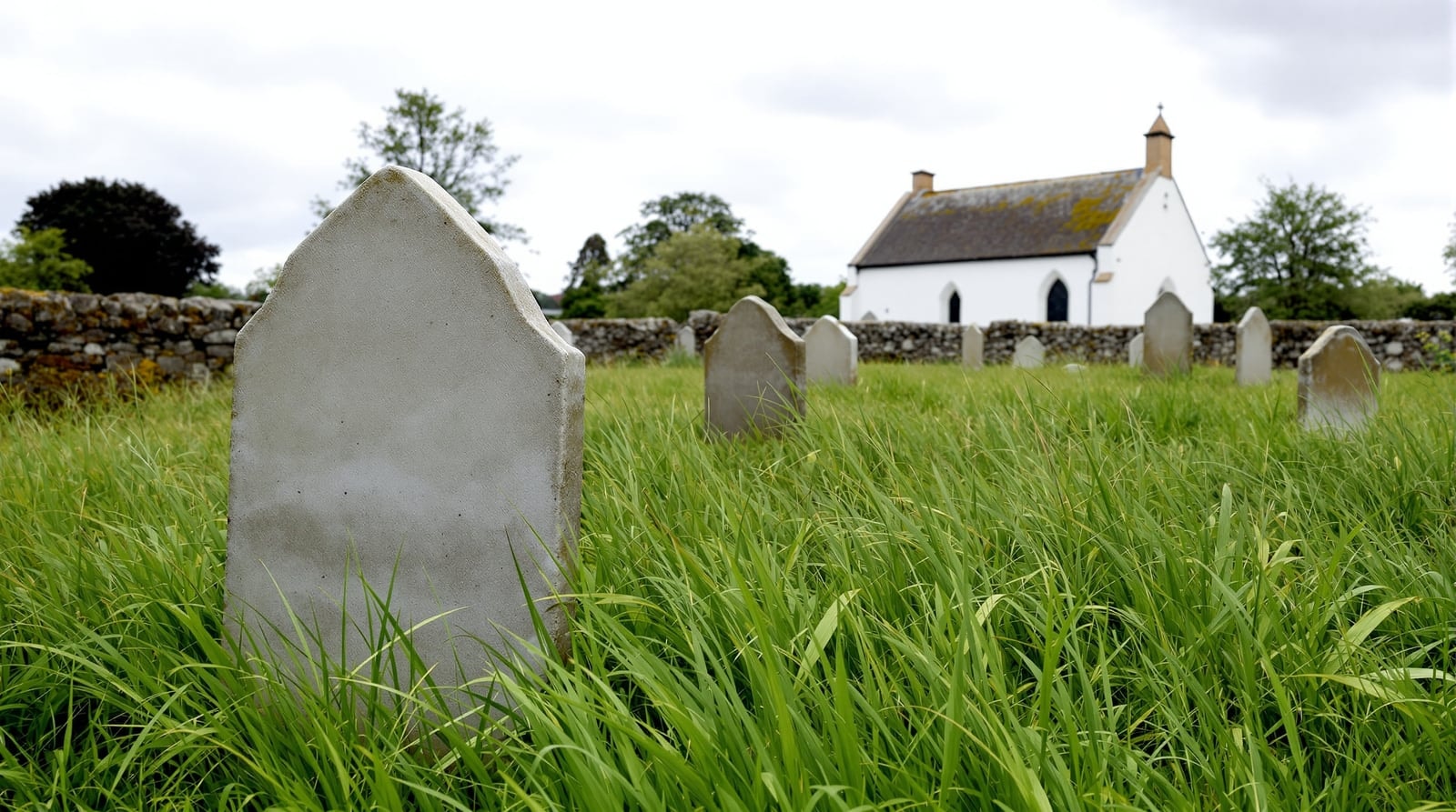 A quiet green churchyard with plain weathered stones in long grass and a small white church beyond a low stone wall