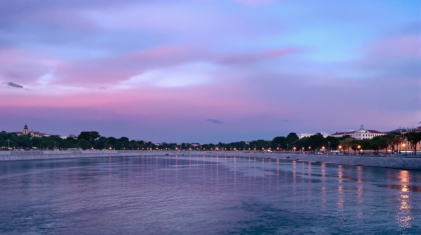 A wide river at dusk with a long stone embankment, glowing lamp posts and a pink and violet sky