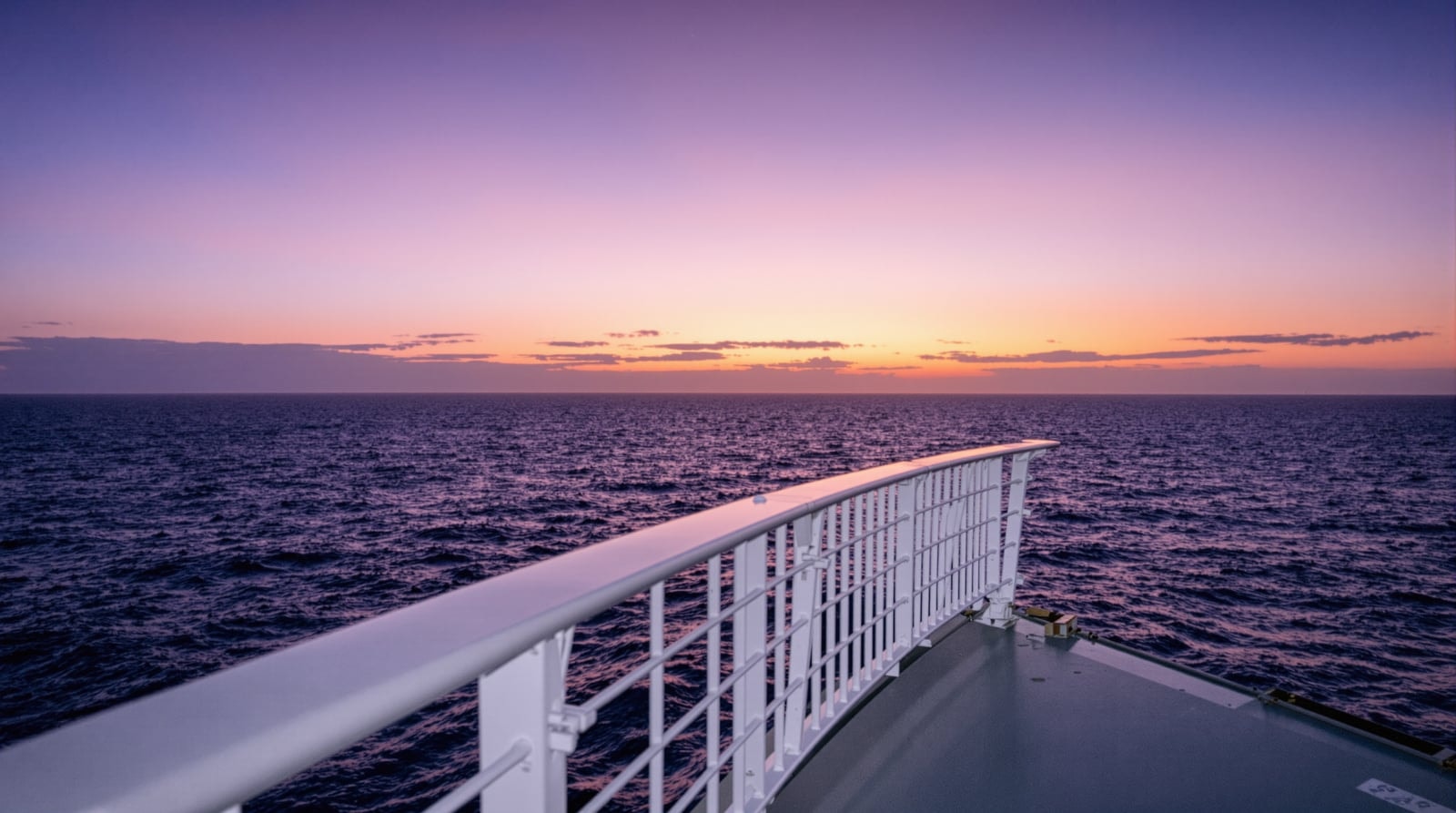 A white ship's deck railing looking out over calm open sea under a violet and orange dusk sky