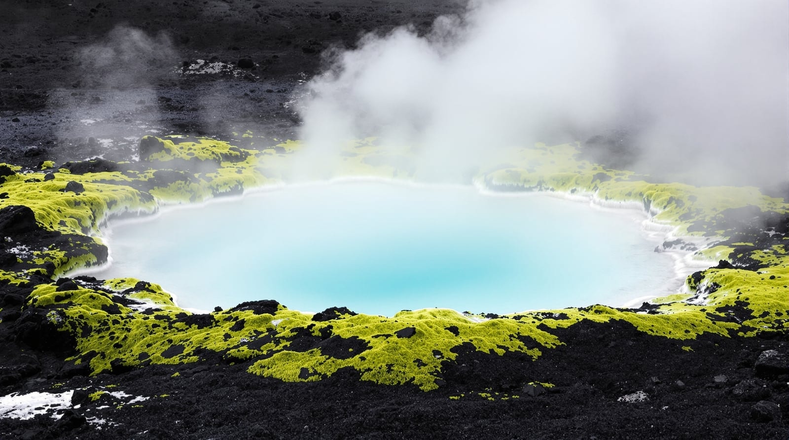 Steam rising from a pale blue natural hot spring pool ringed with green moss in a black lava field